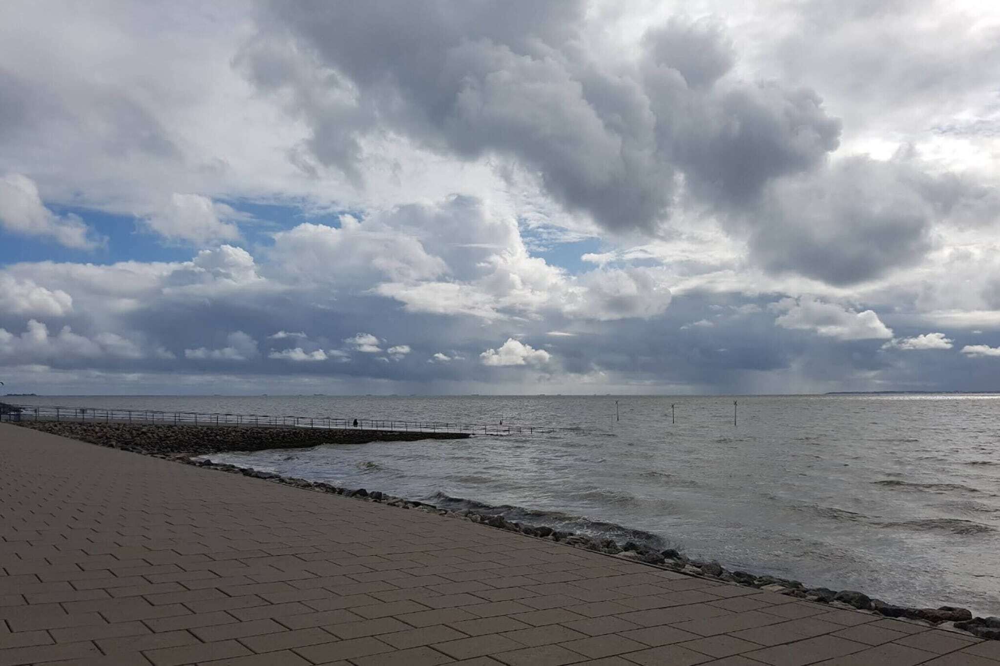 Ein gepflasterter Weg entlang einer felsigen Küstenlinie mit Metallgeländer, der zu einem Gewässer unter einem bewölkten Himmel führt.