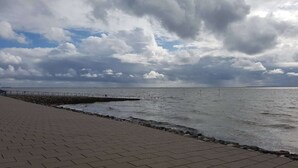 A paved walkway along a rocky shoreline with a metal railing, leading to a body of water under a cloudy sky. - Luxuriös eingerichtetes Ferienhaus in Strandnähe (Dagebüll)