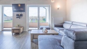 A modern living room with a grey sofa, a coffee table, and a TV mounted on the wall. There is a sliding glass door leading to an outdoor area. - Luxuriös eingerichtetes Ferienhaus in Strandnähe (Dagebüll)
