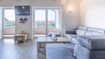 Ein modernes Wohnzimmer mit grauem Sofa, einem Couchtisch und einem an der Wand befestigten Fernseher. Eine Schiebetür aus Glas führt in einen Außenbereich.
