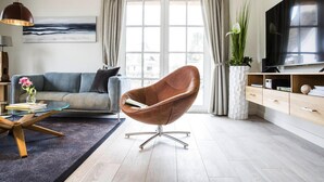 Un salon moderne avec un canapé, une table basse en verre, un fauteuil marron, un téléviseur et une grande fenêtre avec des rideaux.