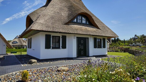 Une maison à toit de chaume, aux murs blancs, aux volets noirs et dotée d’un petit jardin agrémenté de galets et de fleurs.