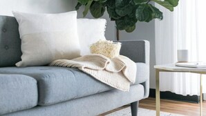 Een grijze bank met een witte kussens, een beige gebreide plaid en een kleine witte schaal op een houten tafel ernaast.