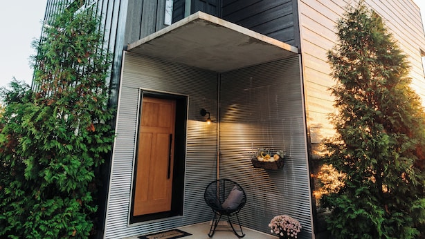 Een modern, twee verdiepingen hoog huis met een schuin dak, een veranda en een kleine tuin met een houten deur en een stoel.