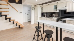 A modern kitchen with white cabinets, a black stove, and a white countertop. There are two black stools and a wooden staircase with black railings.