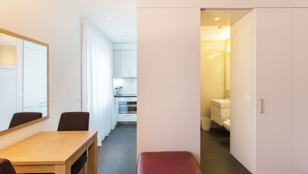 A modern hotel room with a wooden dining table, a red bench, and a view into the kitchen and bathroom. - Nest Wehntalerstrasse (Zürich)