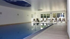 Una piscina con pared de vidrio, rodeada de sillas y mesas, y amplias ventanas con vistas a la vegetación.