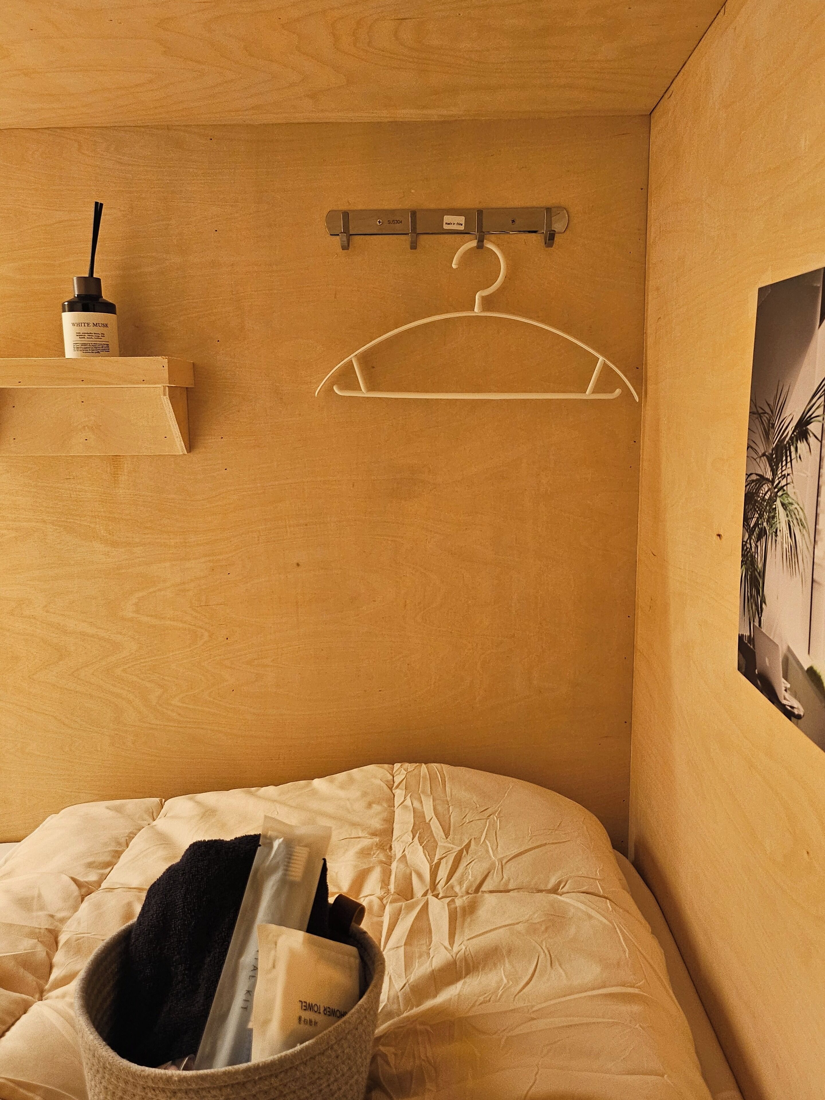 Una habitación de madera con una cama, un perchero, un estante pequeño con una botella y un cuadro en la pared.
