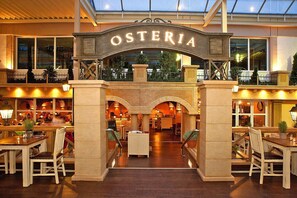 Een hotel lobby met een centrale boog waarop 'OSTERIA' staat, met een bar aan de linkerkant en een zithoek aan de rechterkant.