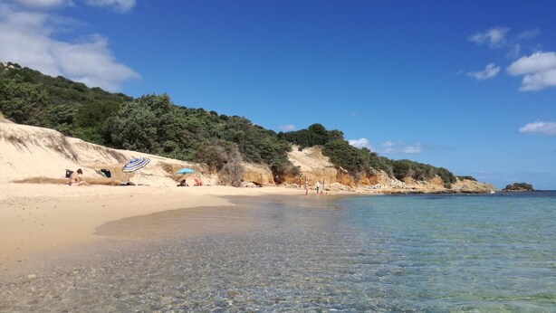 A beach with clear shallow waters, a sandy shore, and a rocky cliff with vegetation. - Apartment in Villaputzu with garden (Chia)