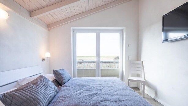 A bedroom with a bed, a chair, a TV, and a balcony with a view. - Exklusiv eingerichtetes Ferienhaus in Strandnähe (Dagebüll)