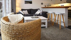 Un salon chaleureux avec un canapé foncé, une table basse blanche et un fauteuil en osier. On aperçoit à l’arrière-plan un coin cuisine avec un tabouret de bar.