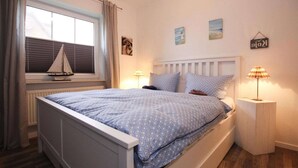 Une chambre à coucher avec un lit, une fenêtre avec des stores, une petite décoration représentant un voilier et une table de chevet avec une lampe.