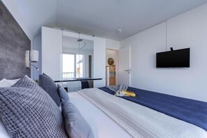 A modern hotel room with a large bed, a TV mounted on the wall, and a view of the city through the window. - Ferienwohnung im Ostseeresort Olpenitz (Ostseeresort Olpenitz)