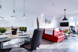 A modern living room with a red sofa, black chairs, a dining table with potted plants, and a large window. - Ferienwohnung im Ostseeresort Olpenitz (Ostseeresort Olpenitz)