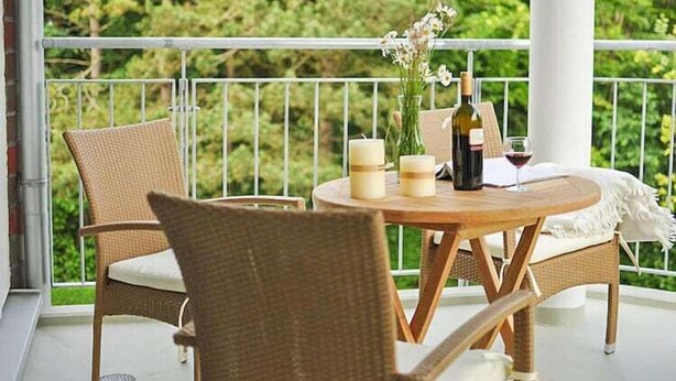A balcony with wicker furniture, a round wooden table, candles, a bottle of wine, and a vase with flowers.