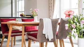 Ein Essbereich mit einem Holztisch, grau und rot gepolsterten Stühlen, einer weißen Vase mit Blumen und Blick ins Freie durch große Fenster.