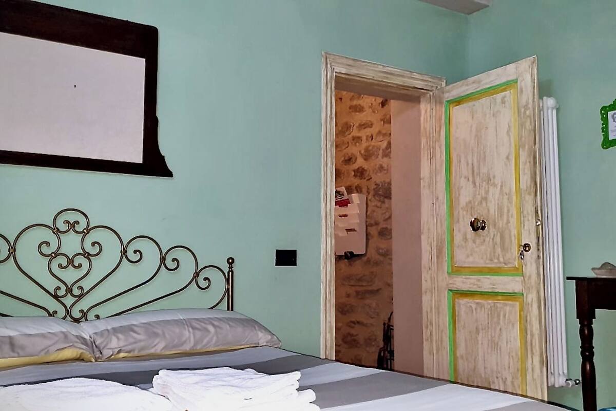 Een slaapkamer met een metalen bed, een nachtkastje met een lamp, en een deur die naar een andere kamer leidt.