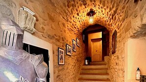 Un couloir aux murs de pierre, avec un escalier menant à une porte en bois. Une personne vêtue d’une armure médiévale se tient sur la gauche.