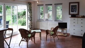 Une pièce spacieuse dotée d’une grande porte-fenêtre coulissante donnant sur un jardin, d’un téléviseur posé sur un meuble et d’une armoire blanche avec des tiroirs.