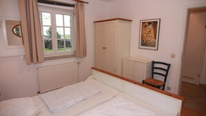 Ein Schlafzimmer mit Bett, einem Fenster mit Vorhängen, einem Heizkörper, einem Kleiderschrank, einem Stuhl und einem Bild an der Wand.