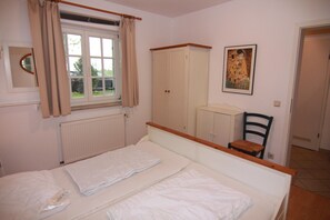 Ein Schlafzimmer mit Bett, einem Fenster mit Vorhängen, einem Heizkörper, einem Kleiderschrank, einem Stuhl und einem Bild an der Wand.