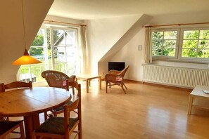 Ein Zimmer mit Esstisch, Stühlen, einem Fernseher und großen Fenstern mit Blick auf Grünflächen.