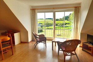 A room with a wooden floor, a dining table, two wicker chairs, a television, and a large window with a view of a green landscape.