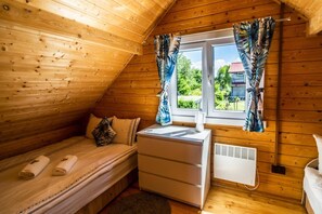 Una habitación pequeña y acogedora con paredes de madera, una cama con colchón y almohadas blancas, una mesita de noche con lámpara y una ventana con cortinas.