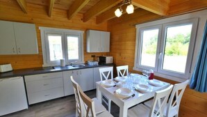Une cabane en bois comprenant une cuisine et un coin repas, avec des meubles blancs, une table à manger pour quatre personnes et de grandes fenêtres laissant entrer la lumière naturelle.