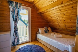 Una cabaña pequeña de una sola habitación con interior de madera, una cama con almohadas y toallas, una ventana con cortinas y suelo de madera.