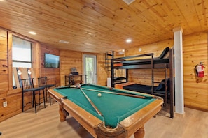 Une table de billard avec des chaises, un téléviseur, des lits superposés et un extincteur dans une pièce en bois.