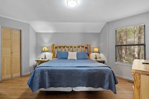 Une chambre à coucher comprenant un lit, une tête de lit en bois, deux tables de chevet avec des lampes, une fenêtre avec des stores et une armoire en bois.