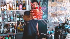 Un barman tient un cocktail décoré de pailles et de garnitures, avec des étagères remplies de bouteilles d’alcool en arrière-plan.