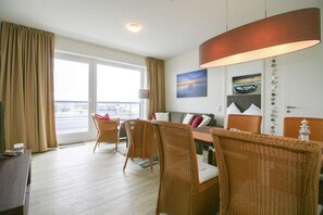 A hotel room with a dining area, wicker chairs, a sofa, a table, and a large window with curtains. - Ferienappartement im Ostseeresort Olpenitz (Ostseeresort Olpenitz)