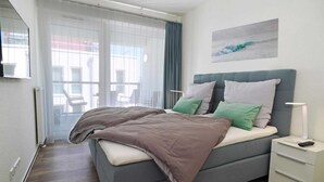 A modern bedroom with a grey bed, white nightstands, and a view through a large window. - Ferienappartement im Ostseeresort Olpenitz (Ostseeresort Olpenitz)