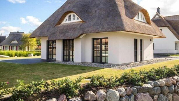 Une petite maison au toit de chaume, dotée de grandes fenêtres et d’une fondation en pierre.