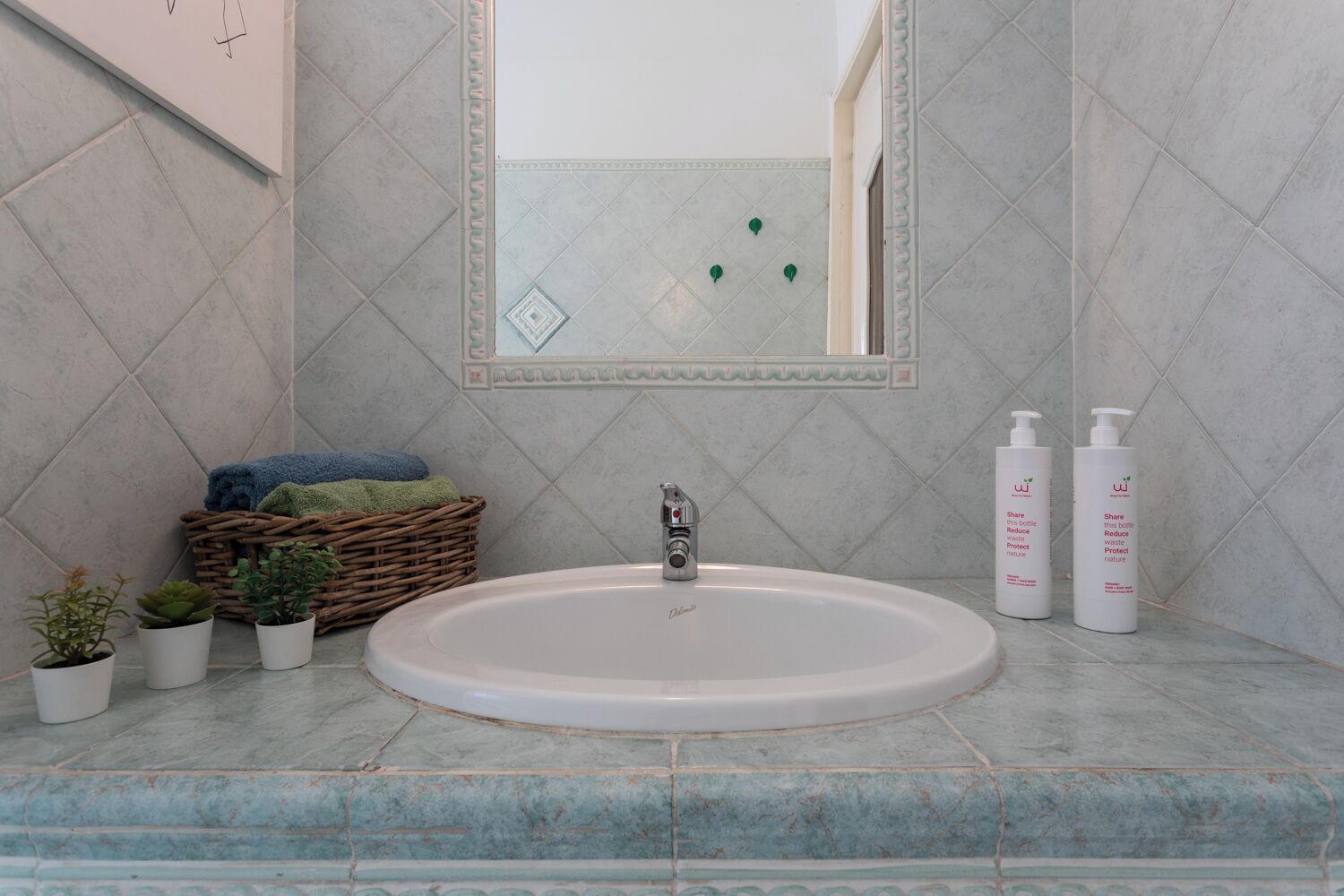 A bathroom with a round sink, a mirror, and two bottles of shampoo. There are also three potted plants and a basket with towels.