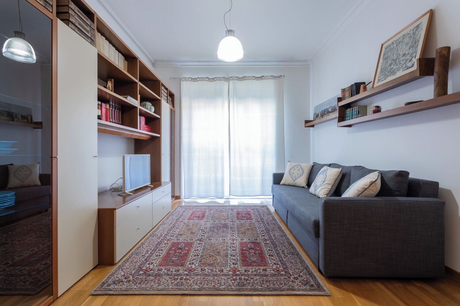 A living room with a grey sofa, a patterned rug, a bookshelf, and a television.