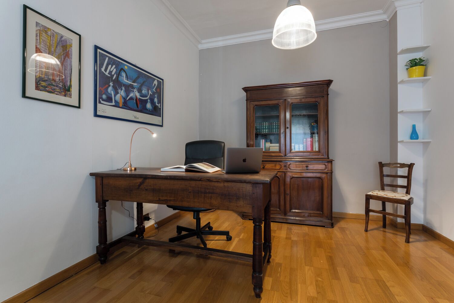 Une pièce comprenant un bureau en bois, une chaise, un ordinateur portable, une lampe, un meuble et deux tableaux accrochés au mur.