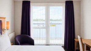 Ein Hotelzimmer mit Bett, Stuhl, Tisch und Blick auf einen Balkon mit Geländer.