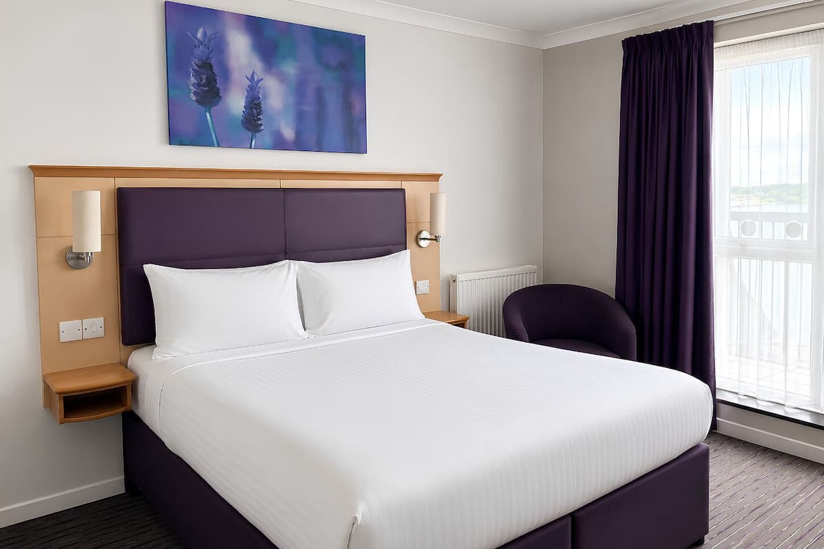 Una habitación de hotel con una cama grande, cabecera, mesita de noche, una silla, una ventana con cortinas y un cuadro en la pared.