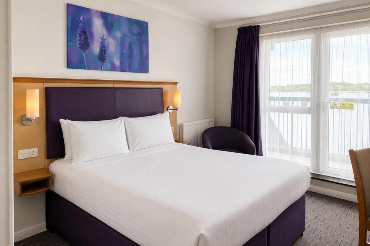 Habitación de hotel con una cama grande, cabecera, mesita de noche, una silla, ventana con cortinas y un cuadro en la pared.