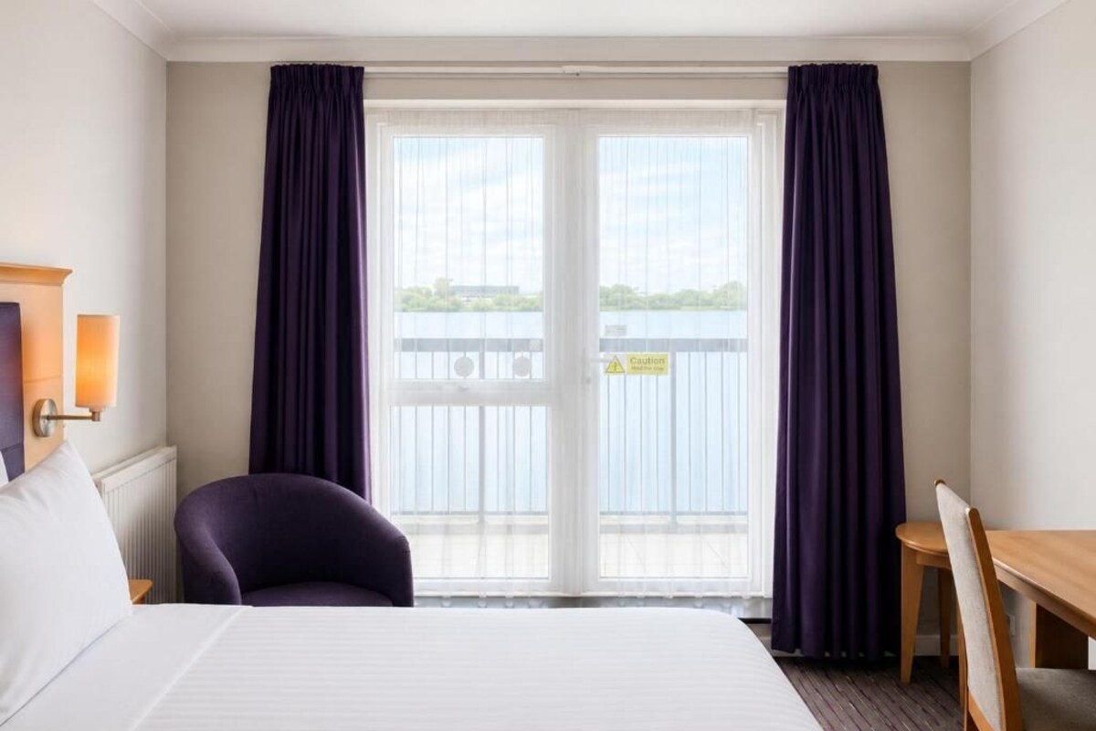 A hotel room with a bed, a chair, a table, and a view of a body of water through a sliding door.