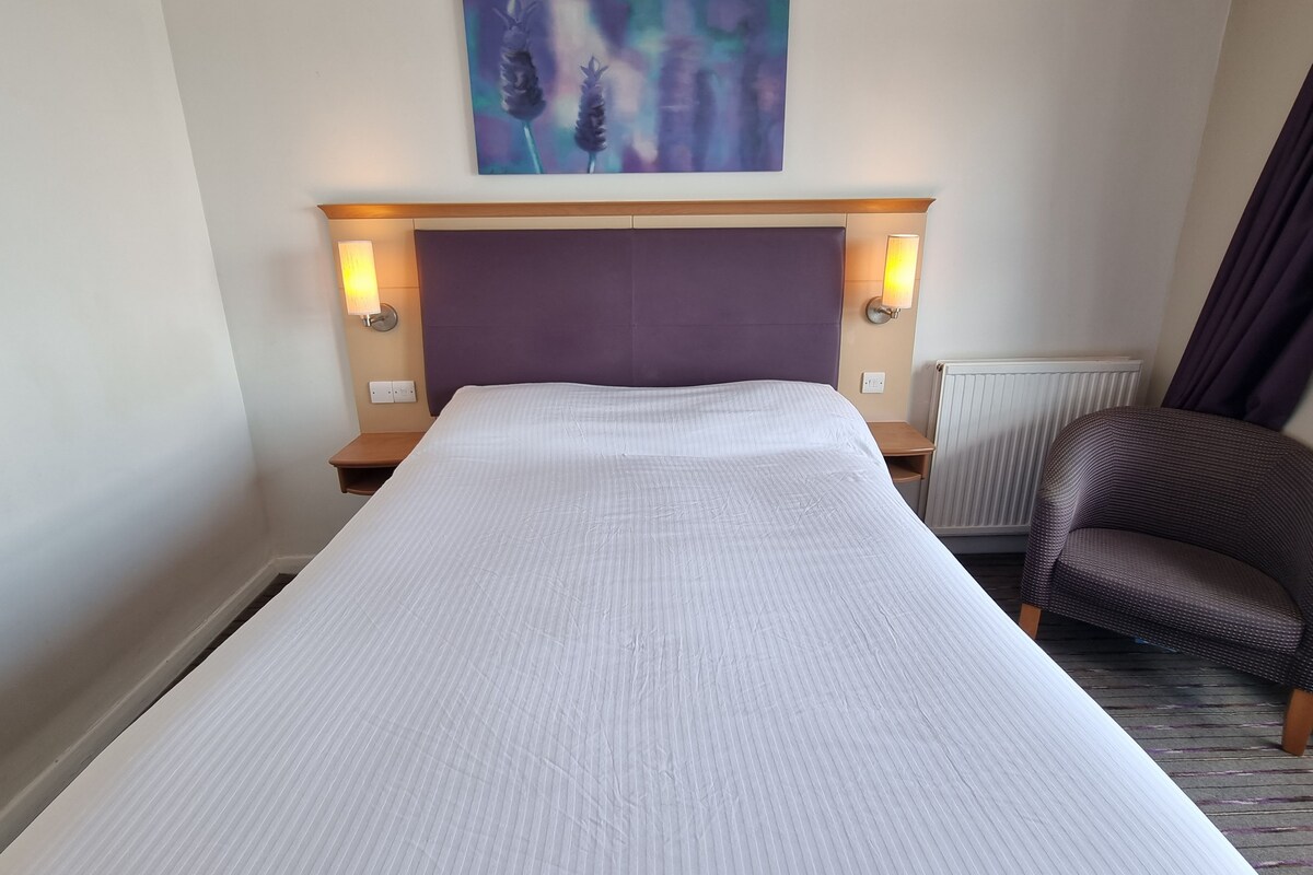 A hotel room with a large bed, a headboard with wall-mounted lamps, a painting above the bed, and a chair.