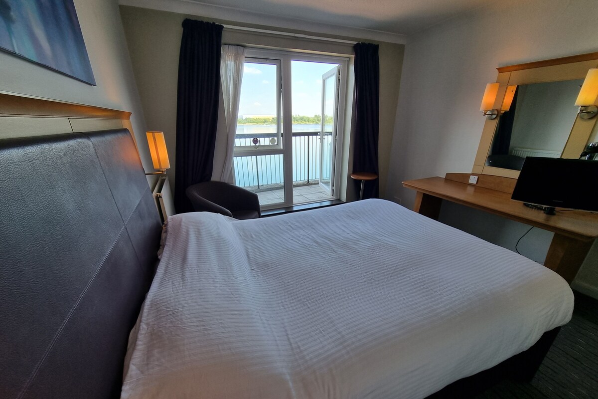 A hotel room with a large bed, a desk with a computer, a chair, and a balcony with a view.