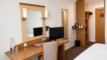 Een hotelkamer met een houten bureau, een stoel, een bed, een televisie, een spiegel, een plank met toiletartikelen en een deur.