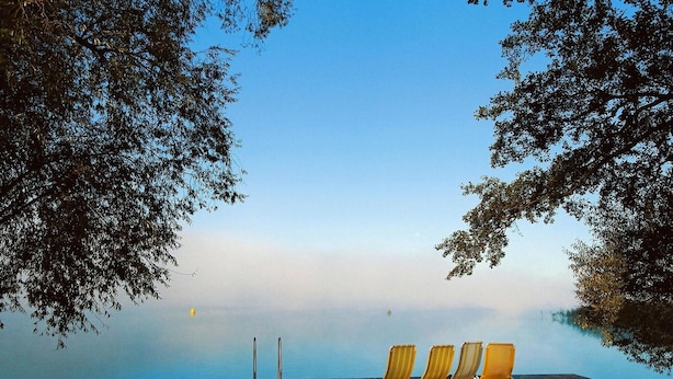 A wooden deck with three lounge chairs facing a calm body of water, surrounded by trees.