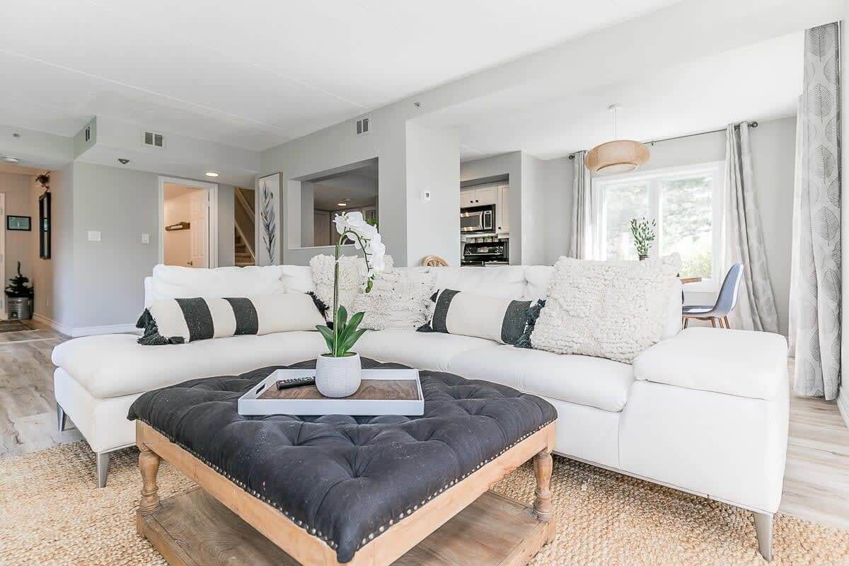 Ein modernes Wohnzimmer mit einem Ecksofa, einem schwarzen Hocker und einer Topfpflanze.