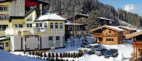 Uma aldeia montanhosa coberta de neve com edifícios de madeira tradicionais, um edifício moderno amarelo e uma vista desobstruída da floresta circundante.